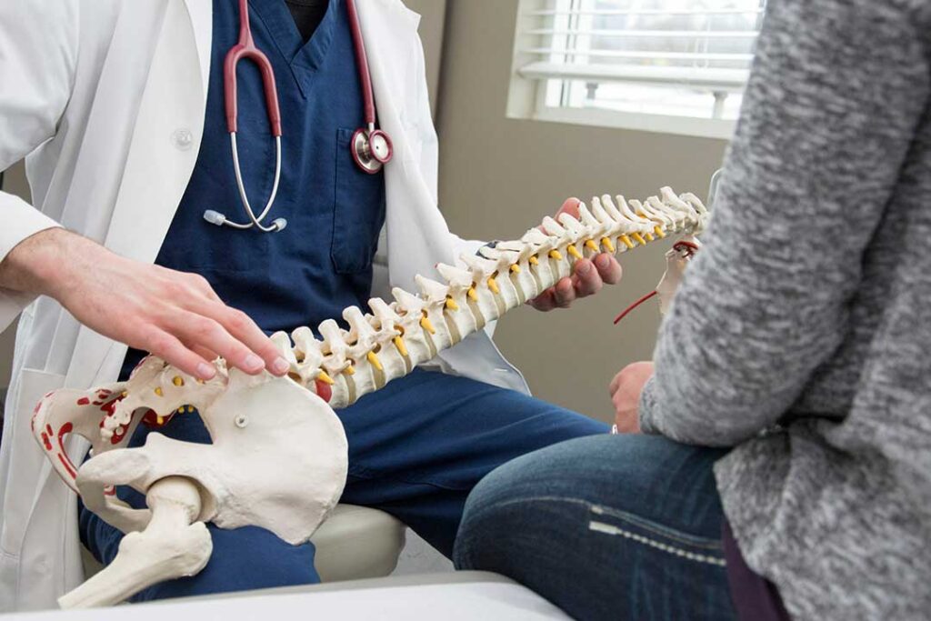 A chiropractor explaining a spine model to a patient in a bright treatment room.