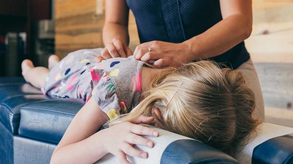 A chiropractor gently checks the posture of a smiling elementary-aged child during a wellness exam.